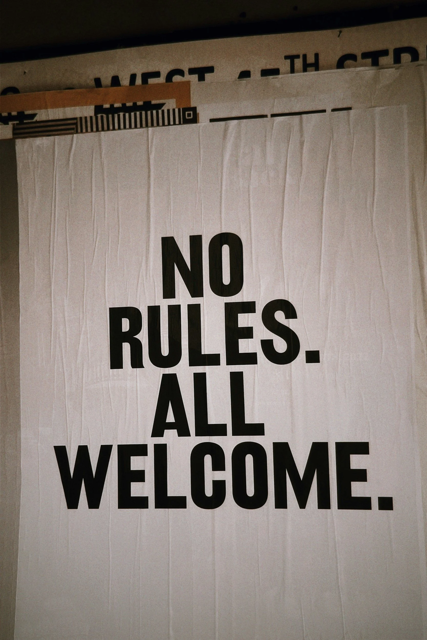 The image shows a white curtain or banner with the large black text 'NO RULES. ALL WELCOME.'