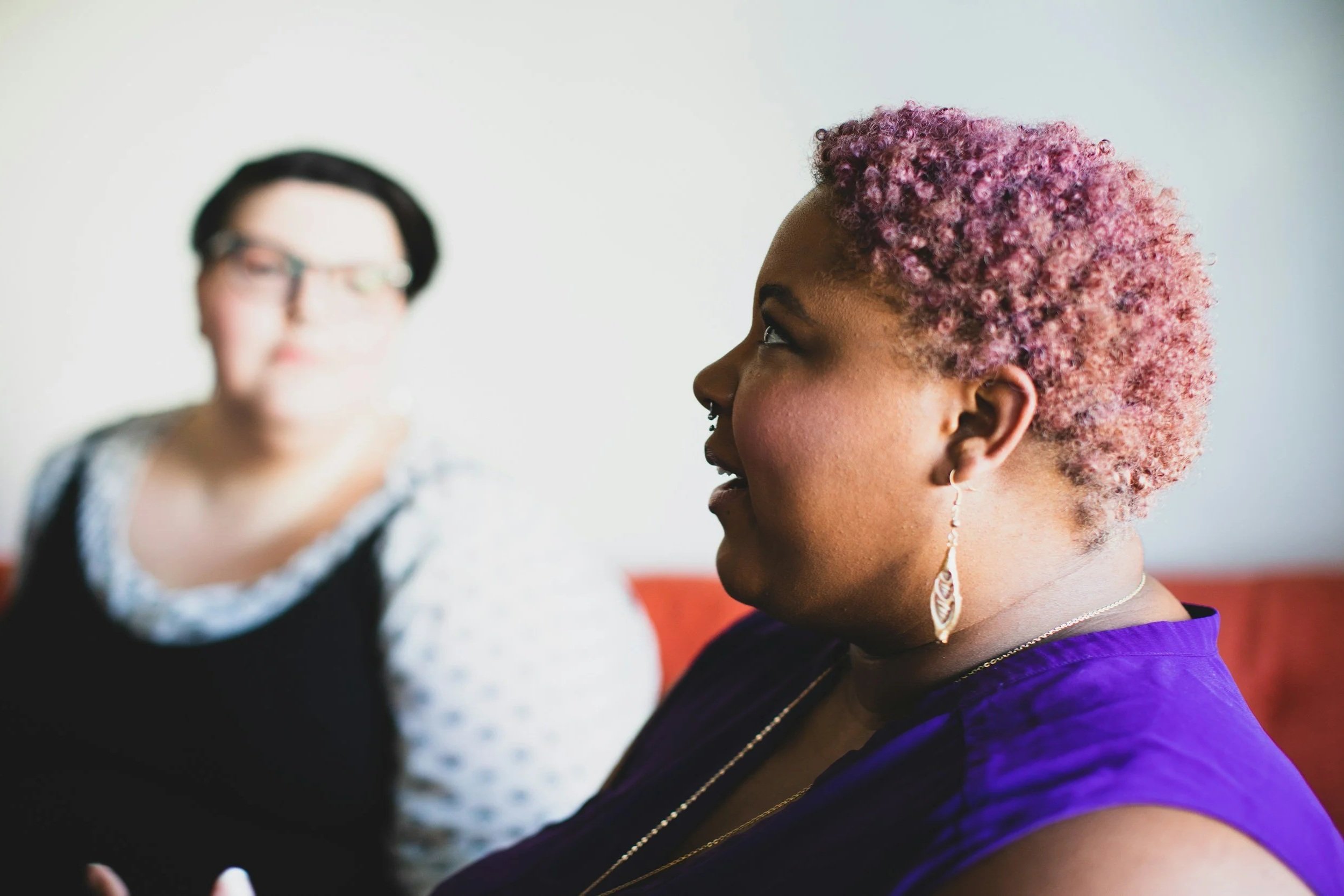 A woman with short, curly, purple-pink hair and earrings, facing to the right, with a second woman blurred in the background, wearing glasses and a black top with white patterned fabric.