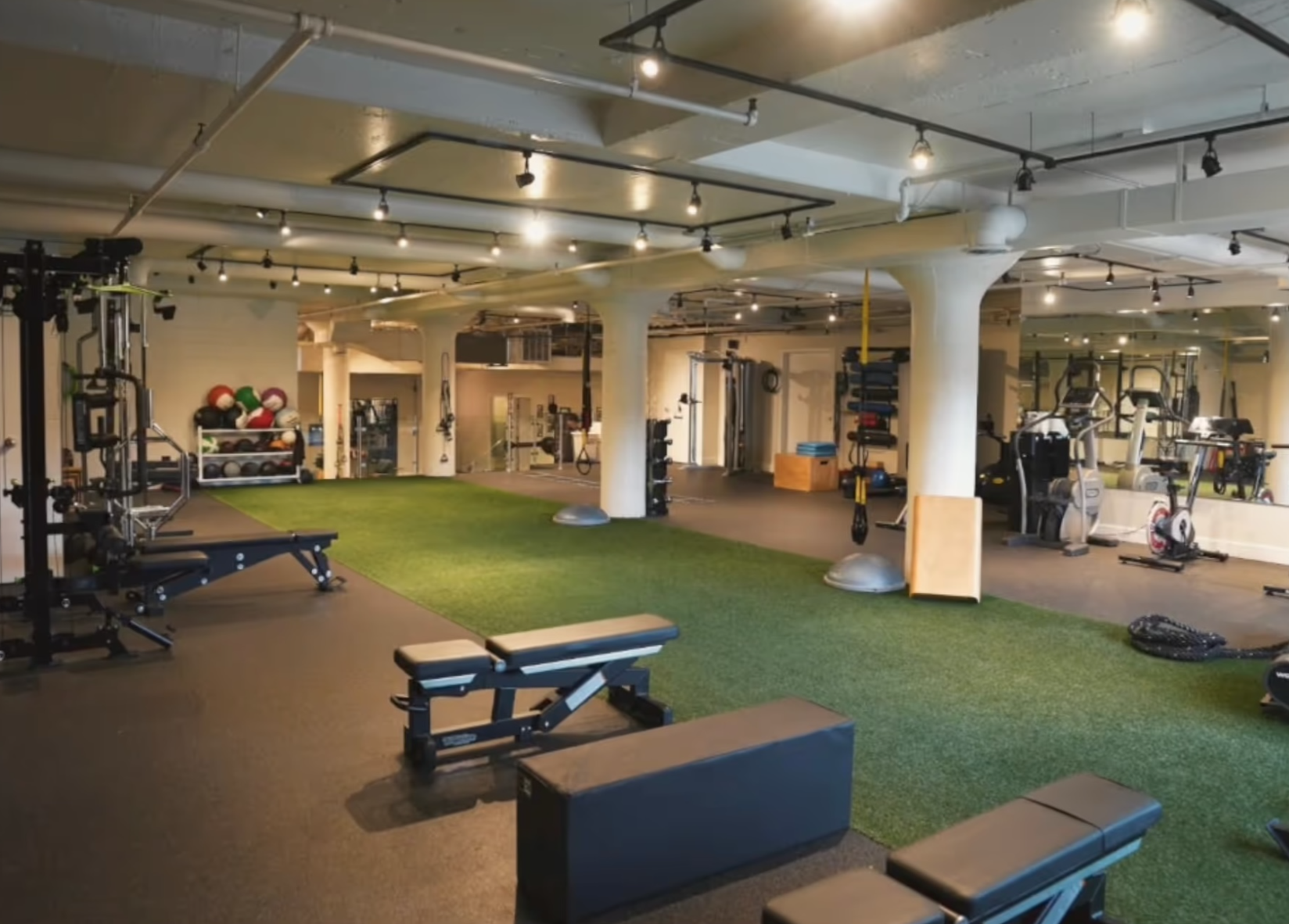 Empty gym with various fitness equipment including weights, exercise bikes, medicine balls, and workout benches, with a green turf area in the center.