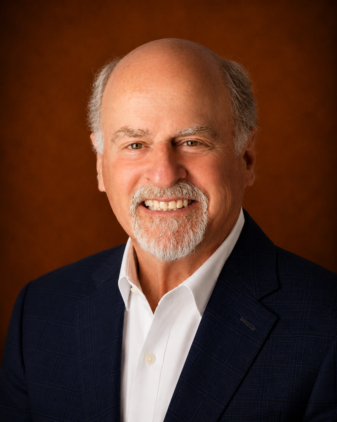 Portrait of an older man with a bald head, gray beard, wearing a white shirt and dark blazer, smiling against a brown background.
