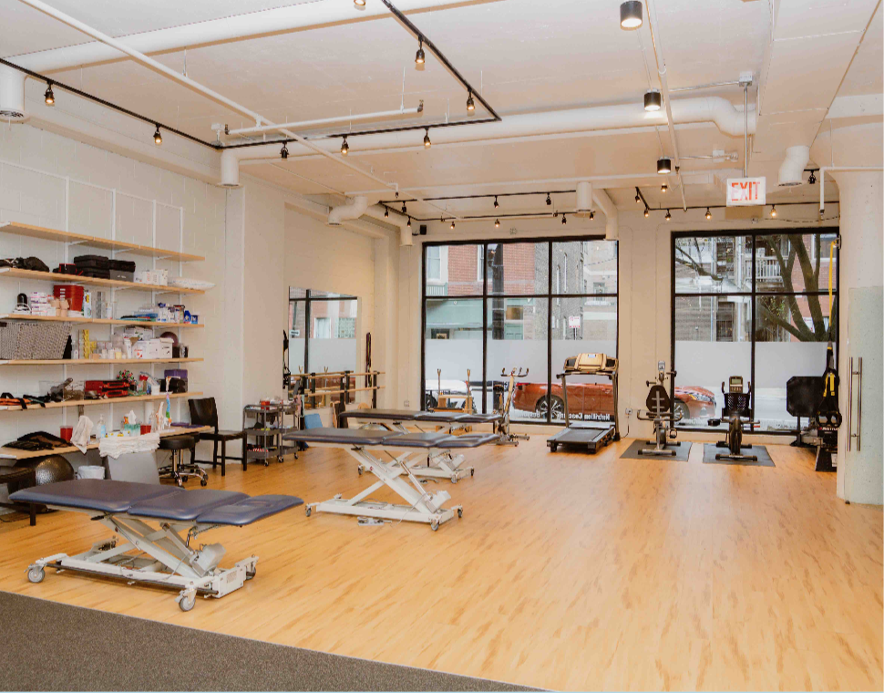 Empty physical therapy or rehabilitation clinic with therapy tables, exercise equipment, and shelves with supplies.