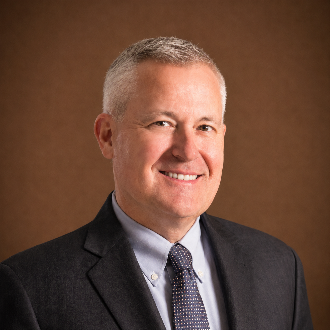 Professional headshot of a smiling middle-aged man with short gray hair, wearing a dark suit, light blue shirt, and a patterned tie, against a brown gradient background.