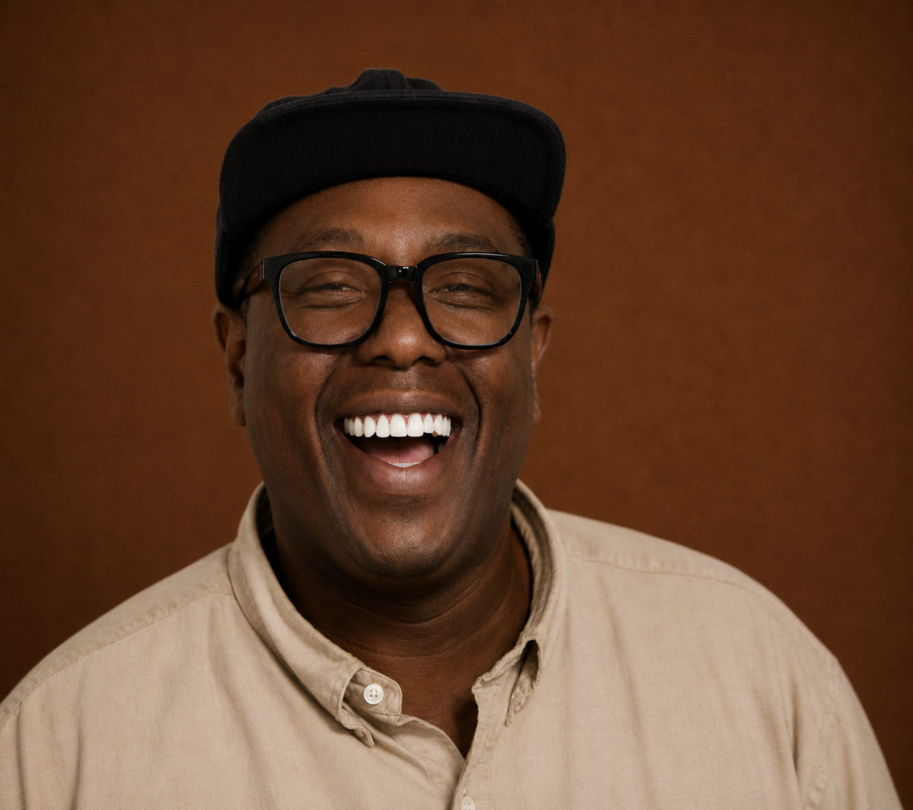 A smiling man wearing glasses, a black cap, and a beige button-up shirt against a brown background.