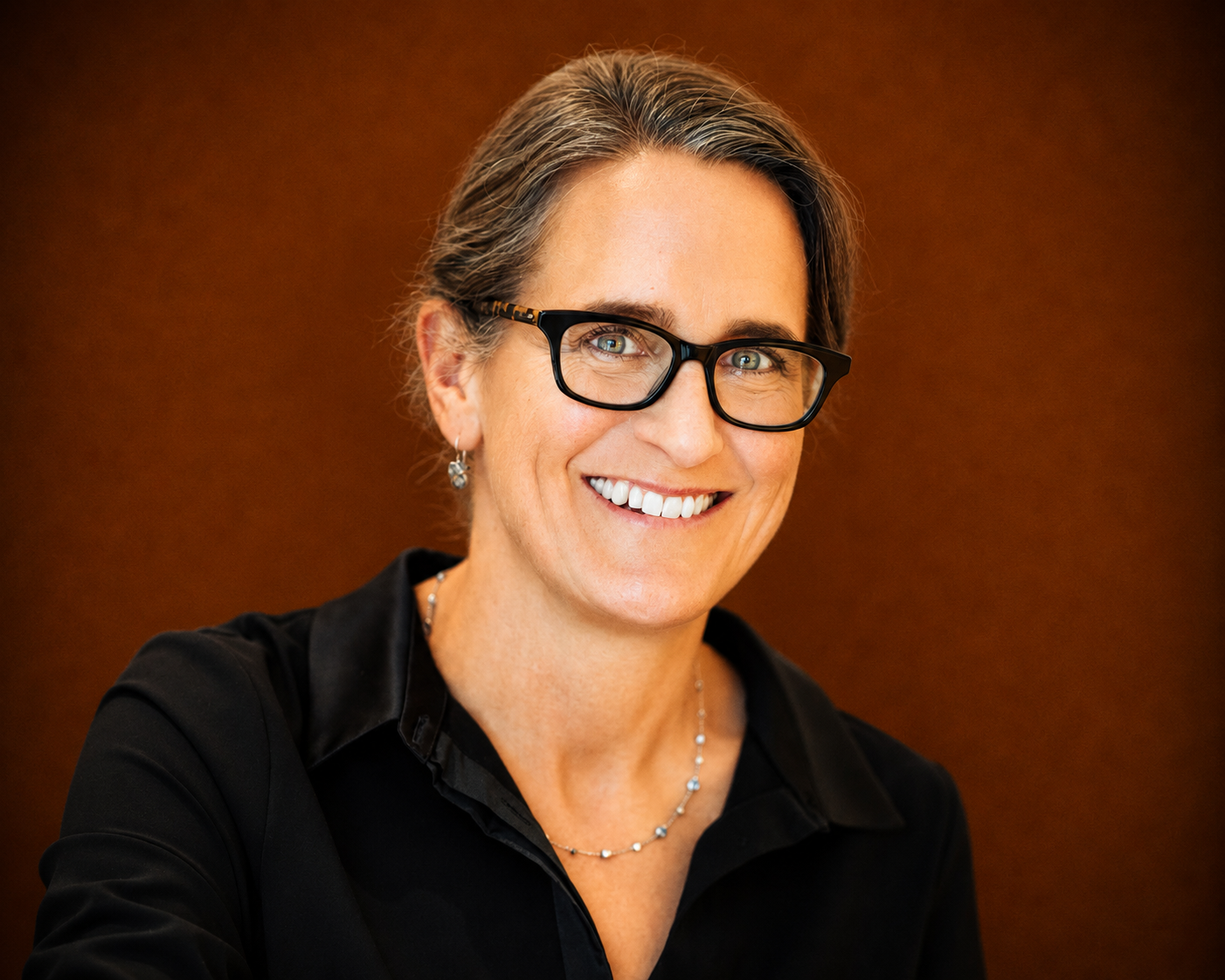 A woman with gray hair styled back, wearing black-rimmed glasses, earrings, a necklace, and a black top, smiling against a brown background.