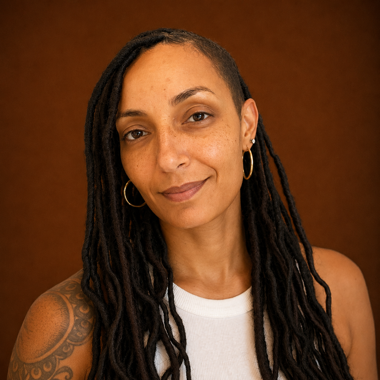 A woman with long dreadlocks, wearing hoop earrings and a white sleeveless top, smiling in front of a brown background.