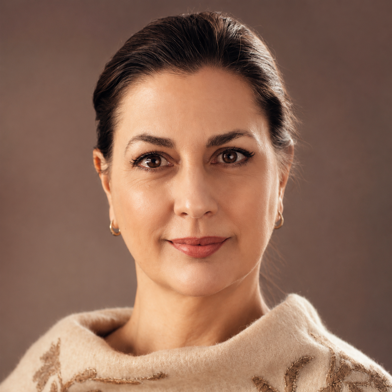 Close-up portrait of a woman with brown hair, wearing gold hoop earrings and a beige shawl with embroidery, smiling softly at the camera.