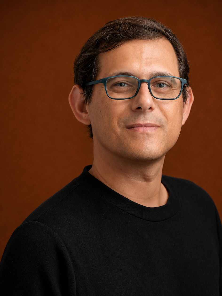 A man with short brown hair, wearing glasses with blue frames and a black shirt, standing against a wooden background.