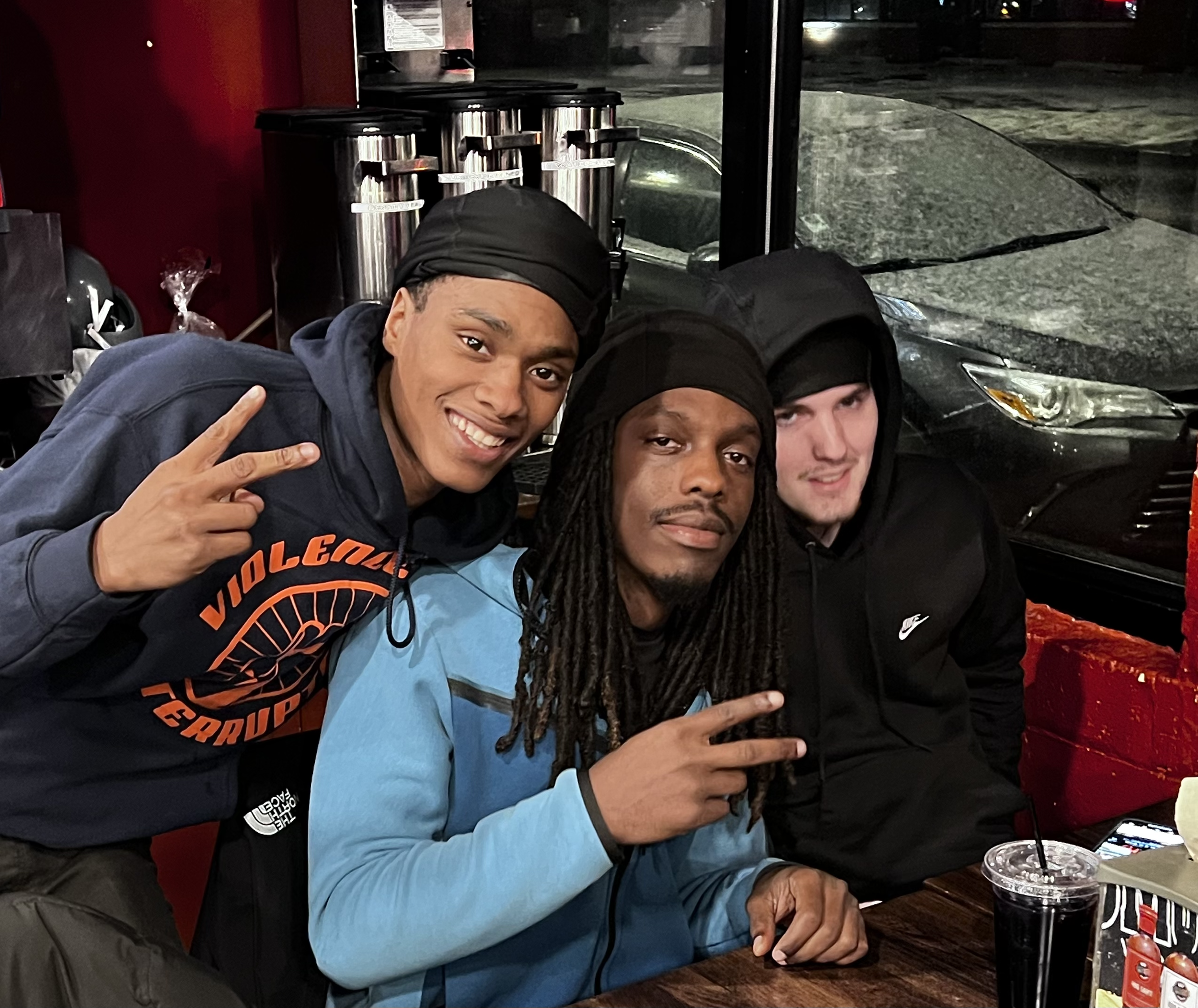 Three young men sitting at a table inside a restaurant or cafe, posing for a photo with peace signs and smiles. Behind them, a window shows a dark street outside, and on the table are drinks and a snack. They are dressed casually, with two wearing hoodies and one with dreadlocks.