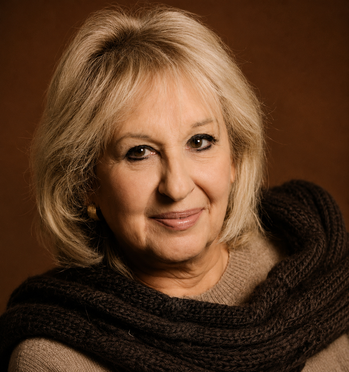 Portrait of an older woman with blonde hair, wearing a beige top and a dark brown scarf, smiling against a brown background.