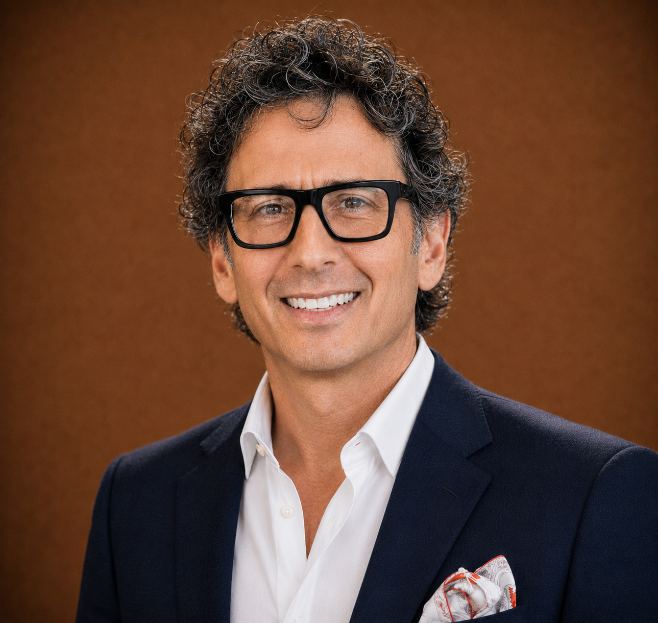 A man with curly dark hair and glasses in a formal suit, smiling against a brown background.
