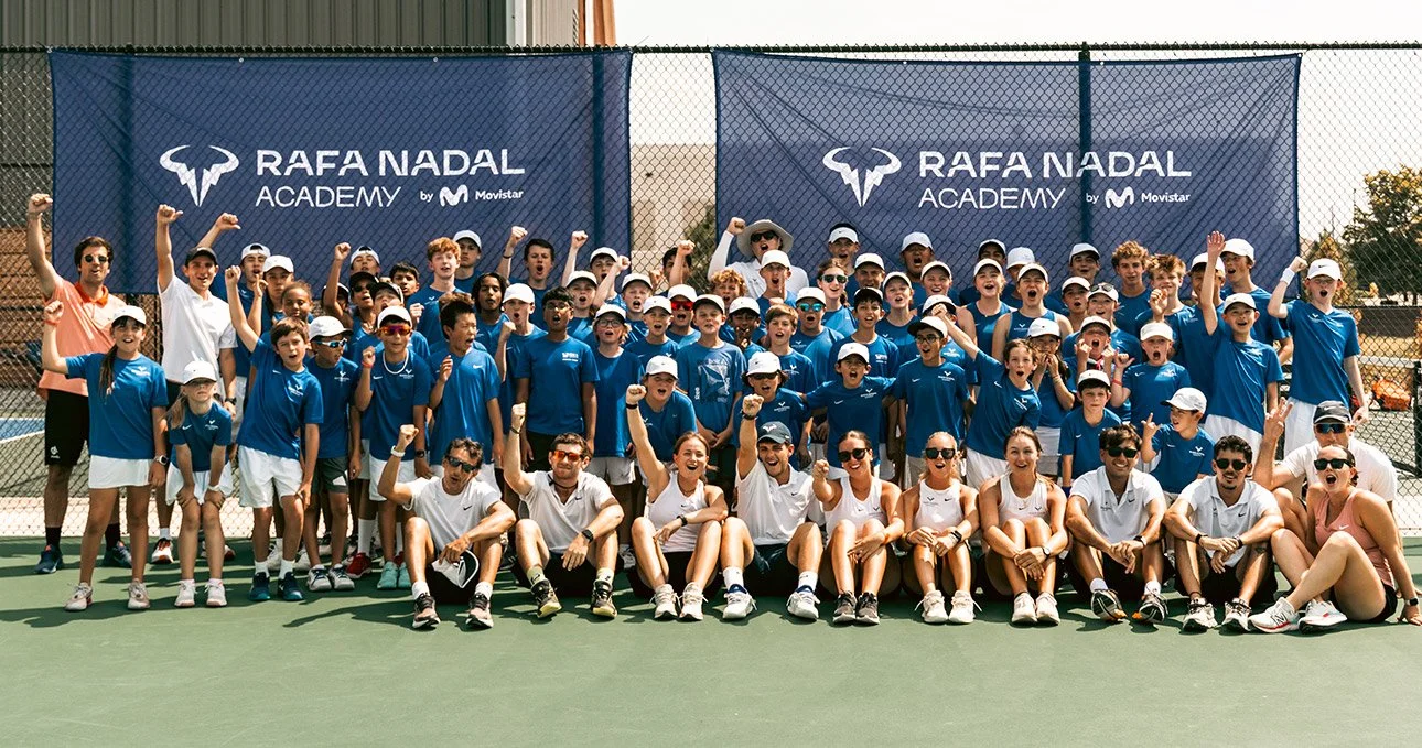 A large group of children and adults posing on a tennis court with a blue banner that reads 'Rafa Nadal Academy by Movistar'. They are celebrating with raised fists and smiling.