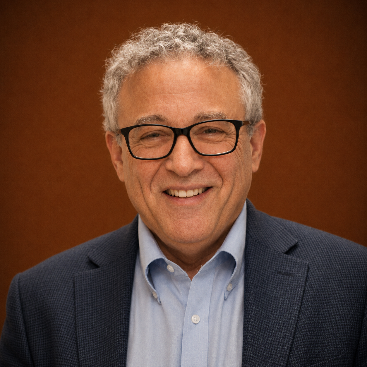 A middle-aged man with curly gray hair, glasses, and a bright smile, wearing a navy blazer and light blue shirt, standing against a brown background.