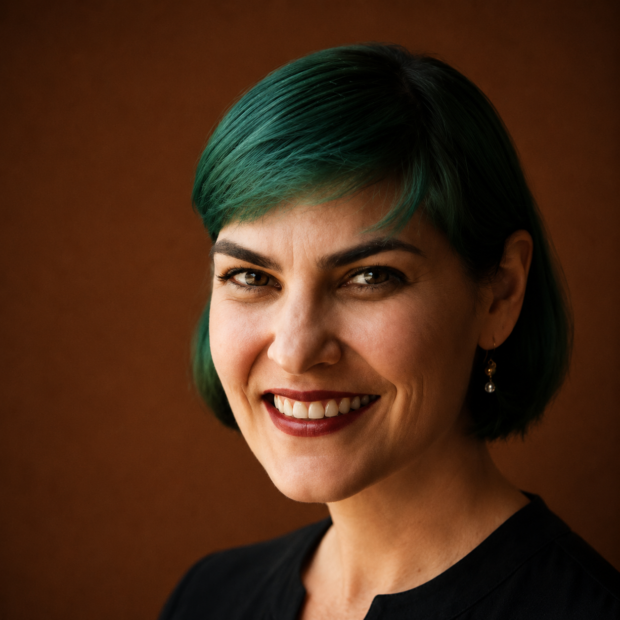 A woman with short, dark hair with green highlights, smiling and wearing a black top and dangling earrings, against a brown background.