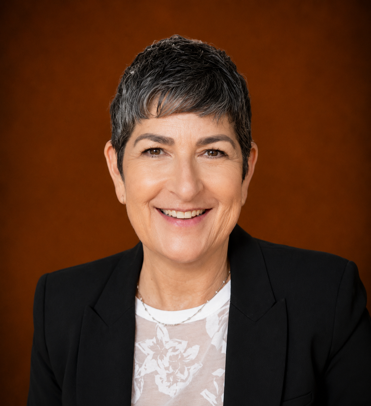 A smiling woman with short gray hair wearing a black blazer and a white patterned shirt, standing against a brown gradient background.