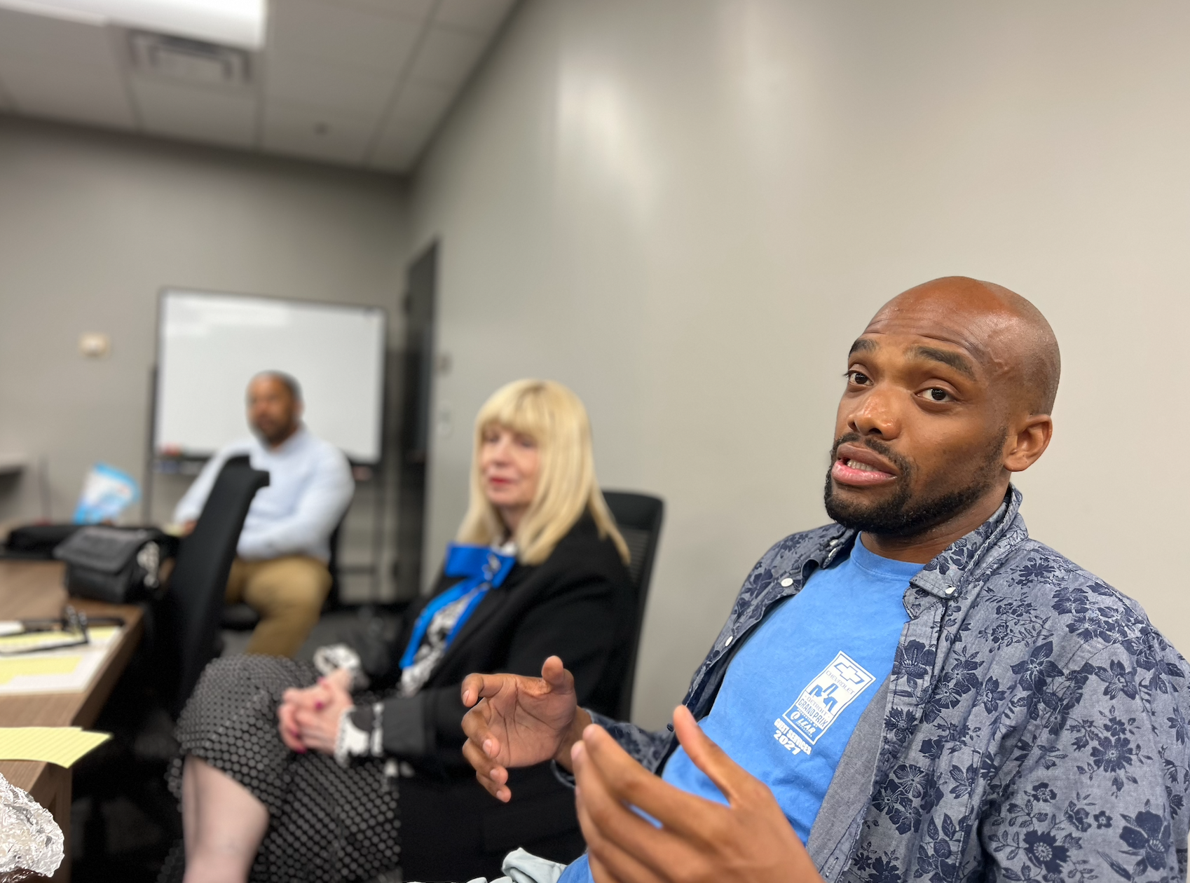 Three people sitting at a conference table in a meeting room. The man in the foreground is looking at the camera with a slight smile, wearing a blue shirt and a floral-patterned jacket. In the background, a woman with blonde hair and a man with dark skin are seated, blurred, in discussion.