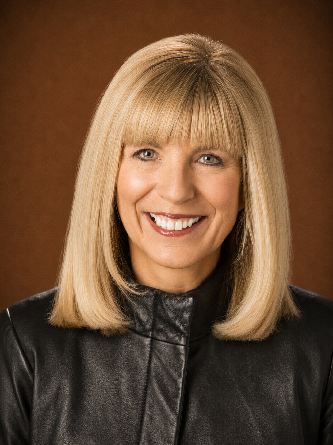 A woman with blonde hair, wearing a black leather jacket, smiling against a brown background.