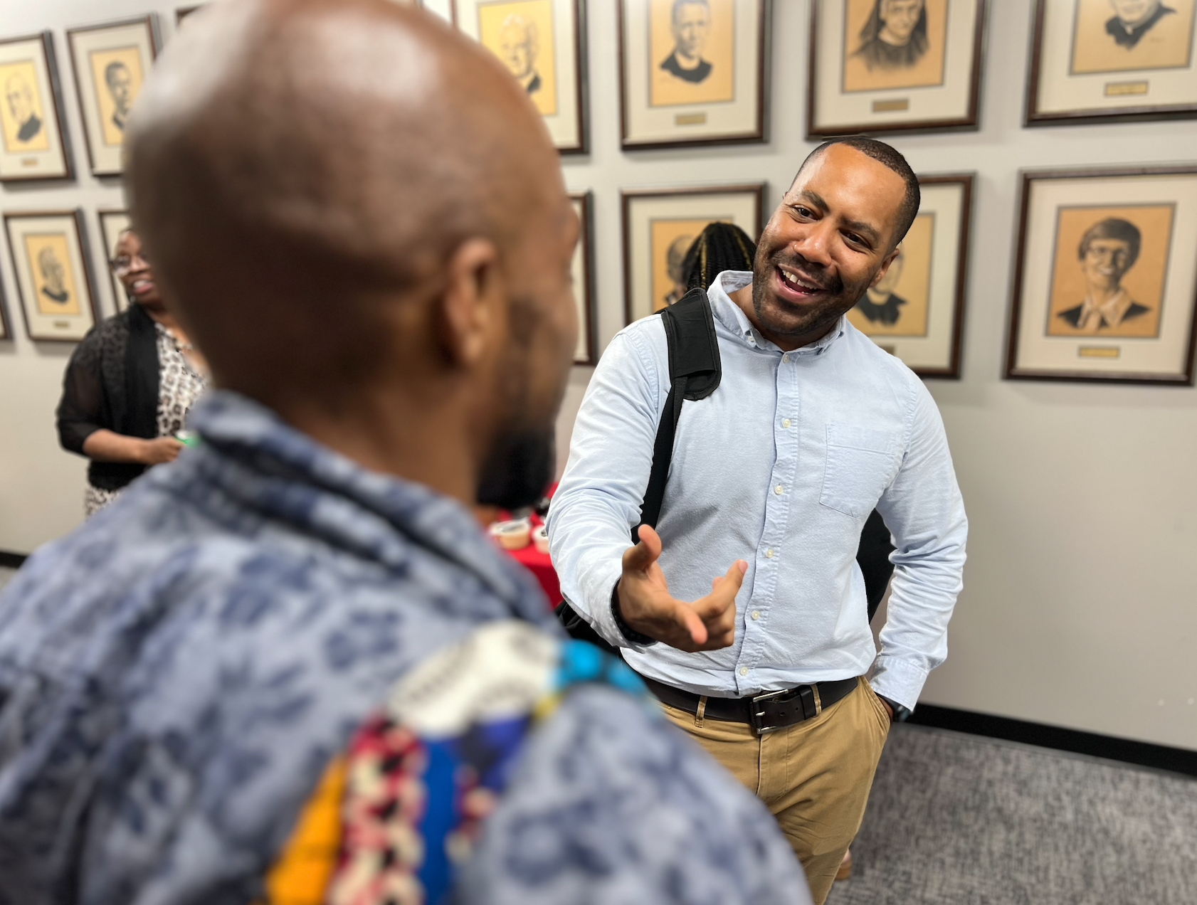 Two men engaged in conversation in an indoor setting with framed portraits on the wall behind them. One man is smiling and gesturing with his hand, wearing a light blue button-up shirt and khaki pants. The other man has his back to the camera, with a bald head, glasses, and a patterned shirt. A woman in the background is smiling and wearing a black and white patterned dress.