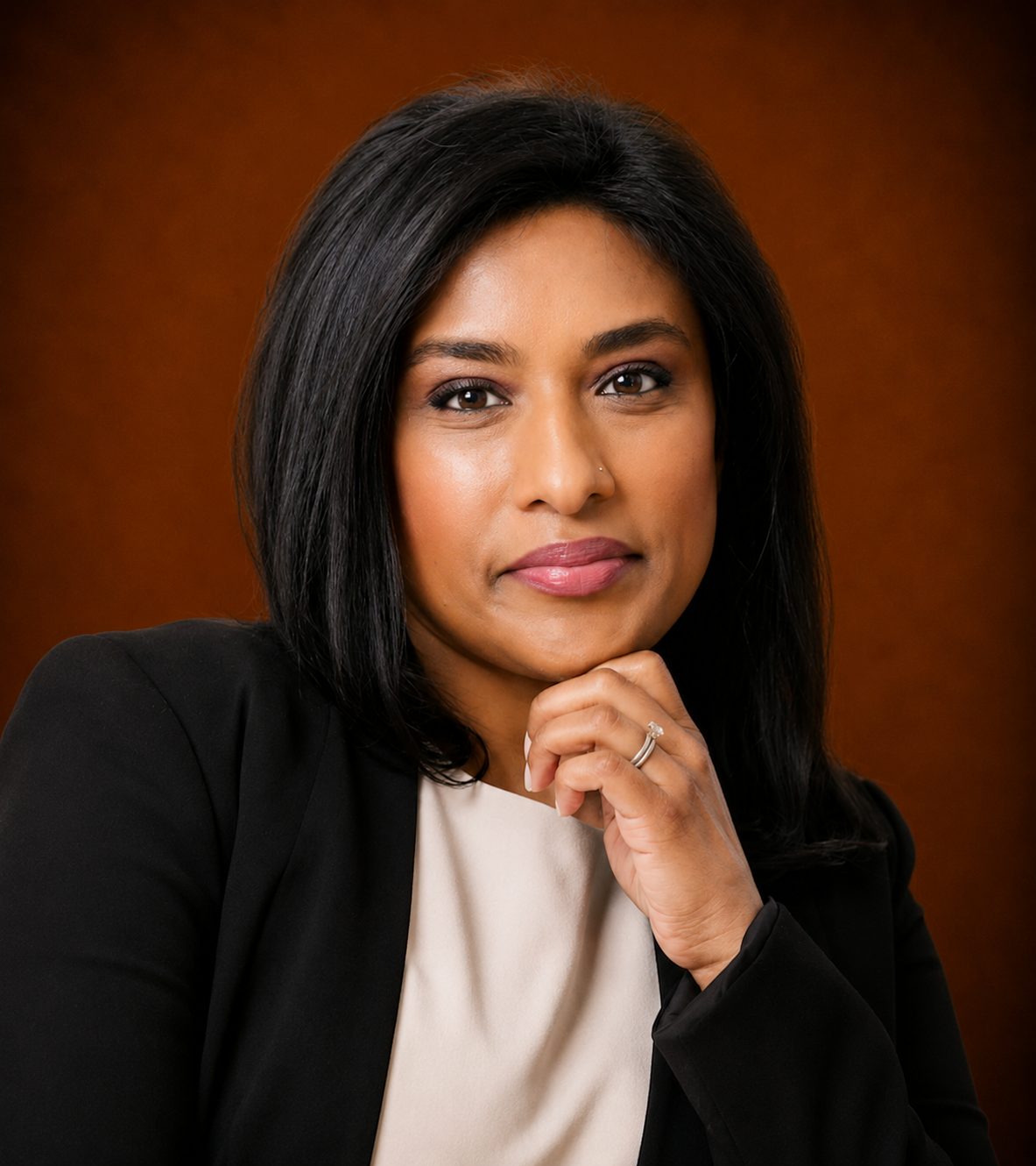 A professional woman with black hair, wearing a black blazer and light-colored top, resting her chin on her hand, against a brown background.