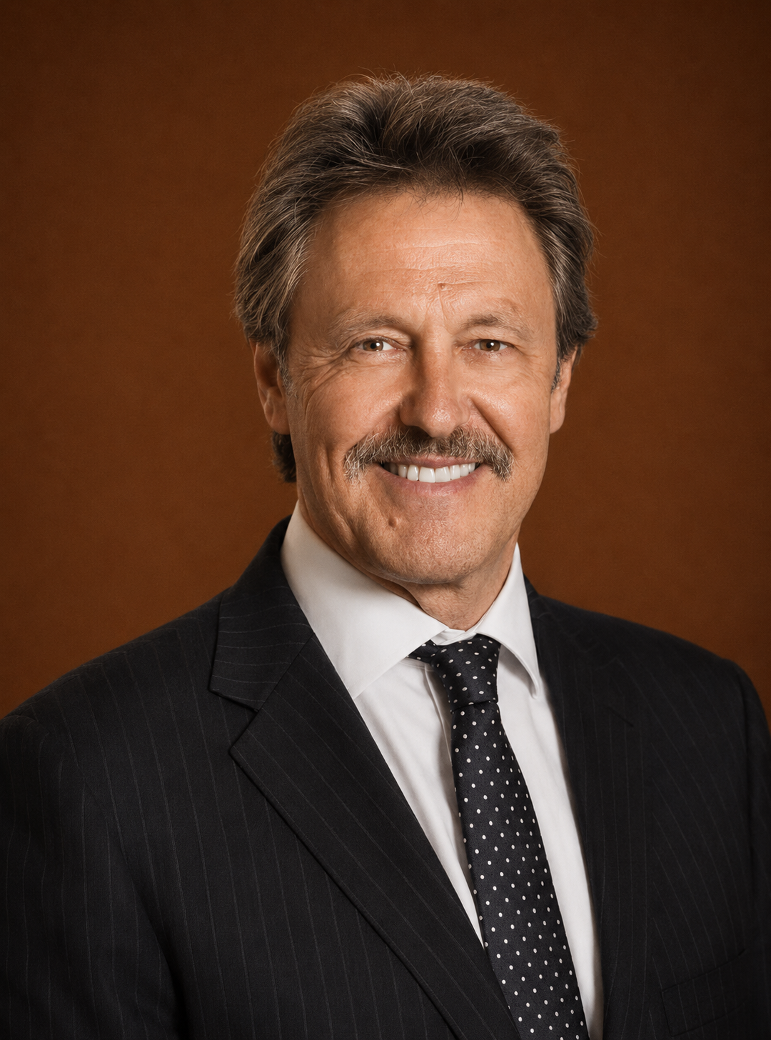 A middle-aged man with gray hair and a mustache, wearing a dark pinstripe suit, white shirt, and polka dot tie, smiling against a brown background.