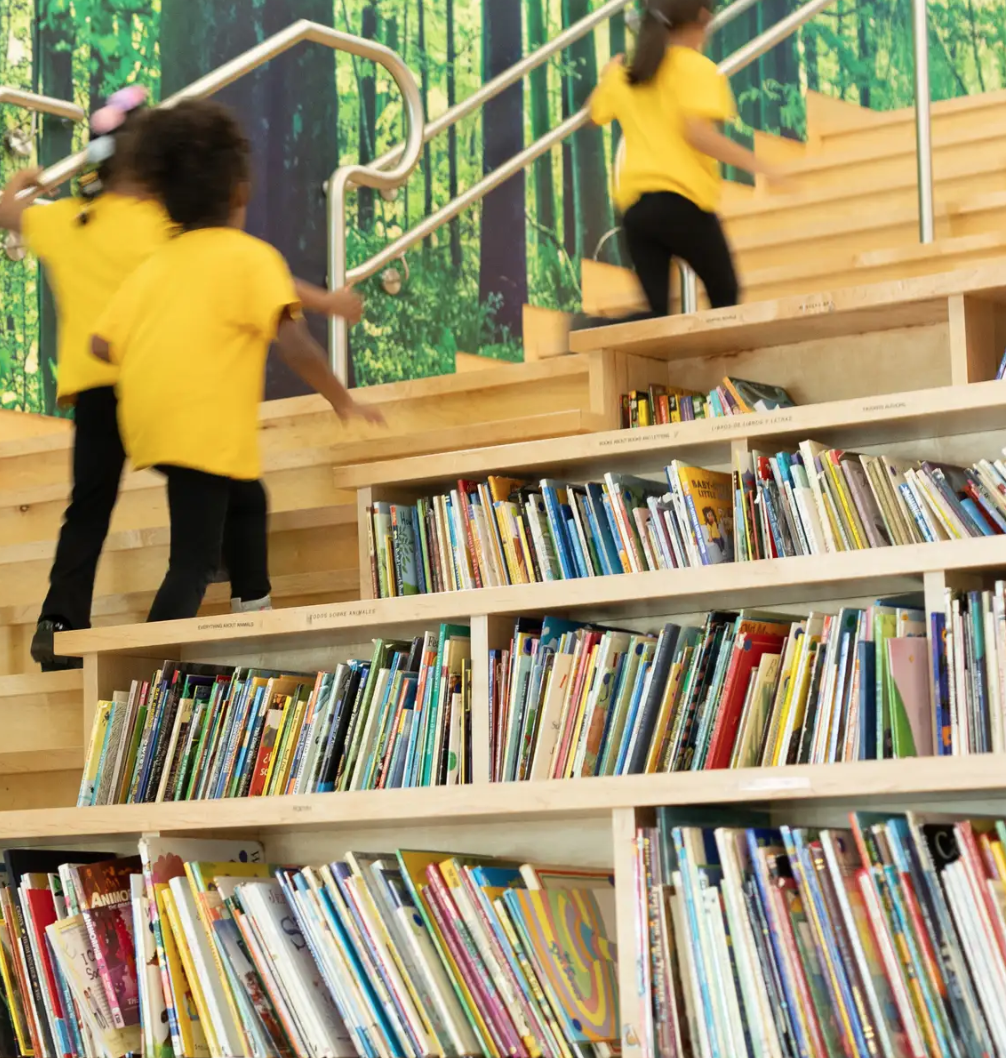 Children in yellow shirts climbing wooden stairs in a library with a forest mural background and bookshelves filled with colorful books.