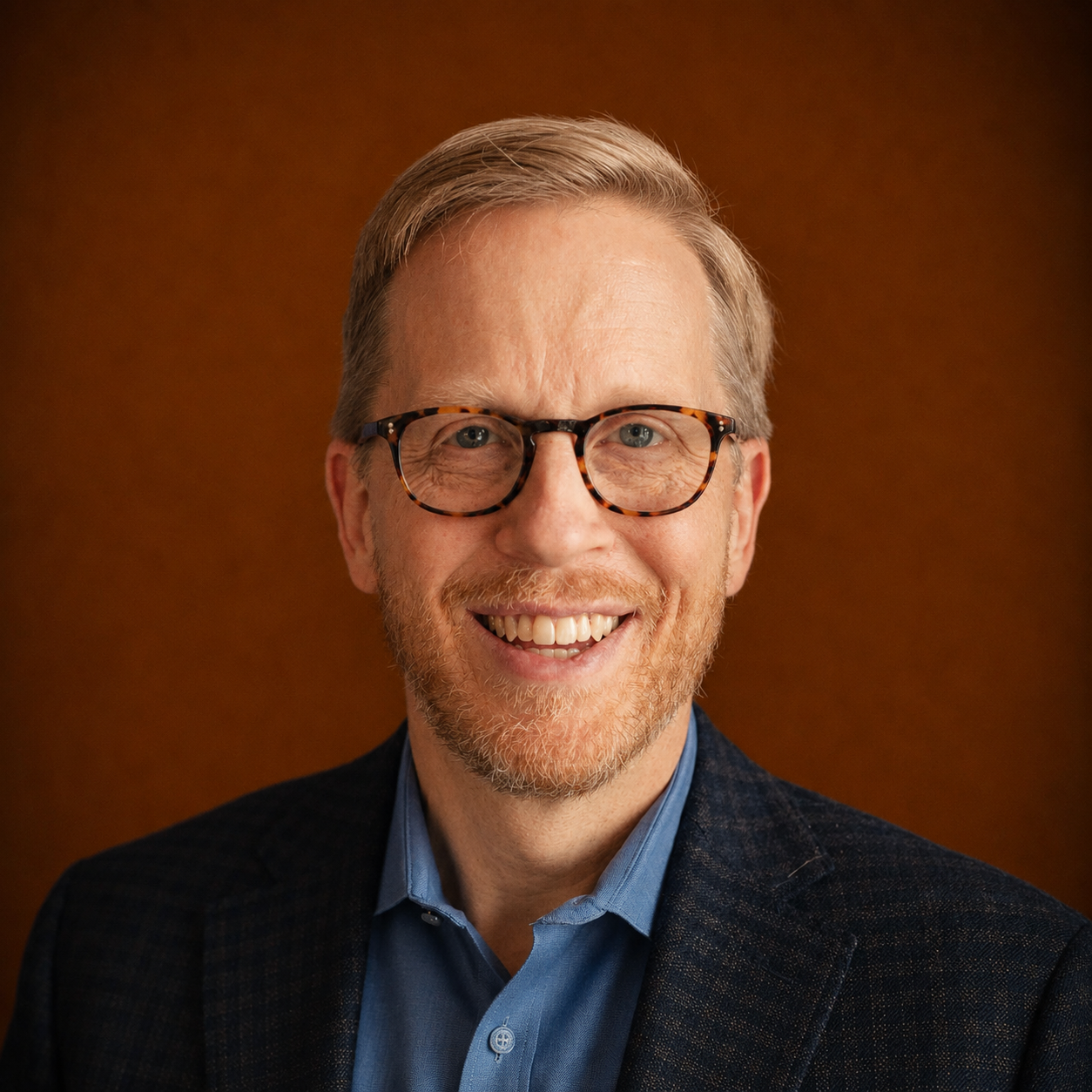 Smiling man with glasses, light hair, beard, wearing a blue shirt and dark blazer, against a brown background.