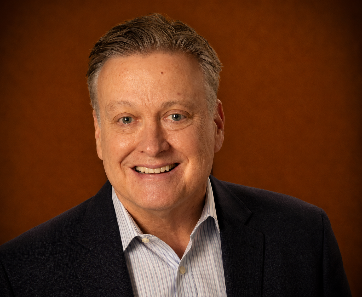 Headshot of a smiling middle-aged man with short, light-colored hair, wearing a dark blazer over a striped dress shirt, set against a brown background.