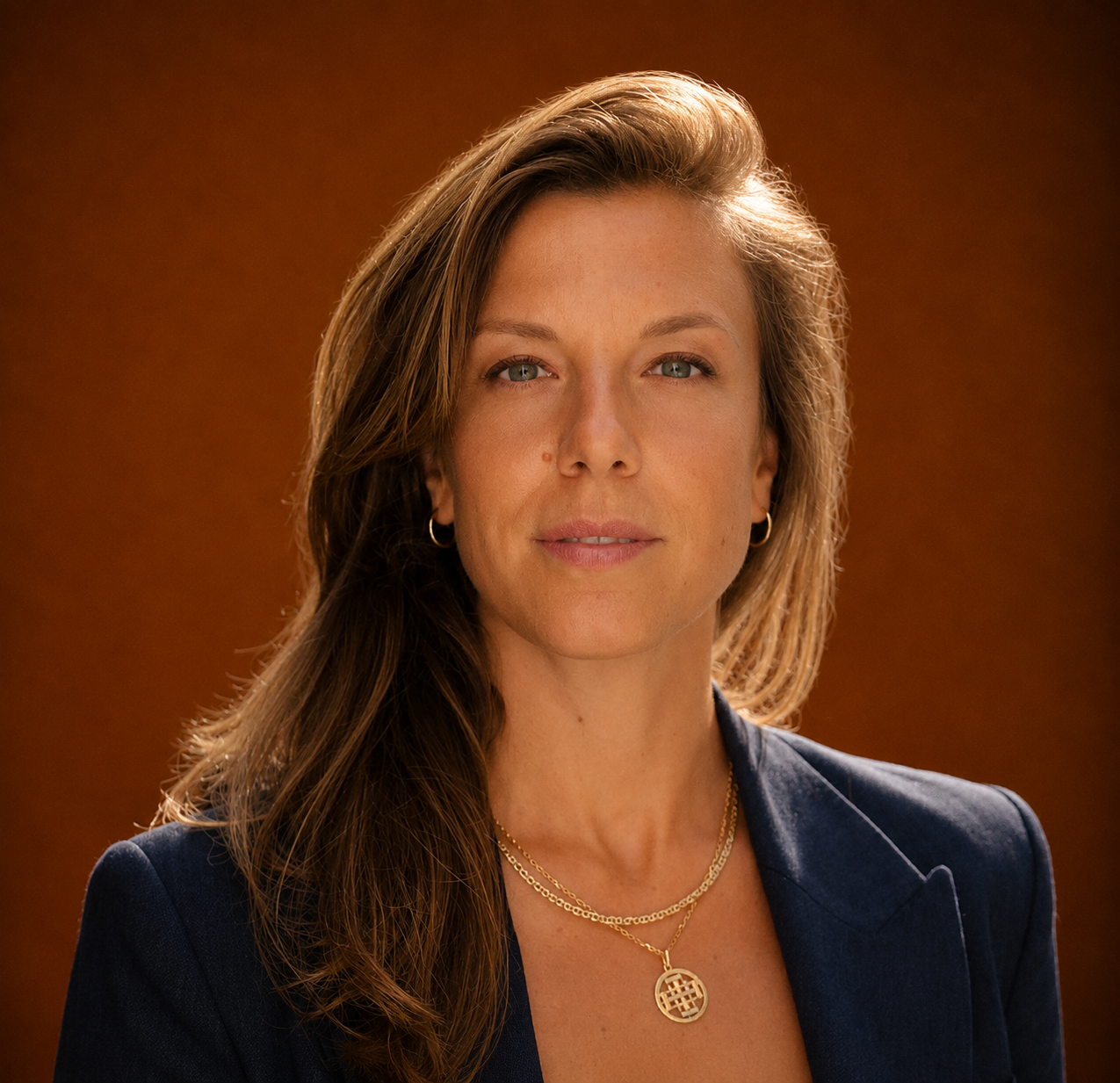 A woman with long brown hair wearing a navy blazer and gold jewelry, looking directly at the camera with a neutral expression, against a brown background.