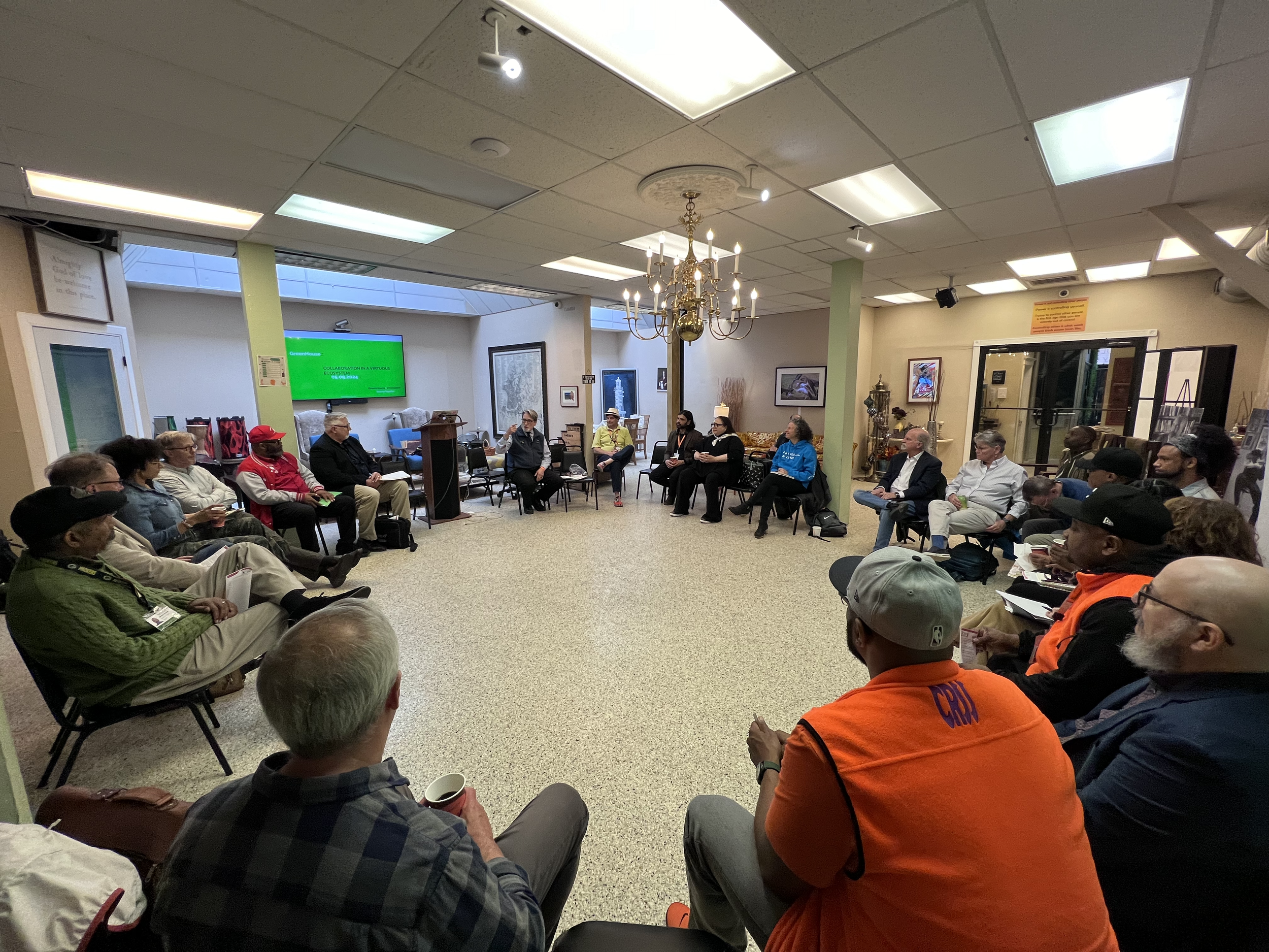 A group of diverse people sitting in a circle attending a meeting or discussion in a conference room. There are artworks on the walls, a chandelier hanging from the ceiling, and a large screen displaying a green slide with text.