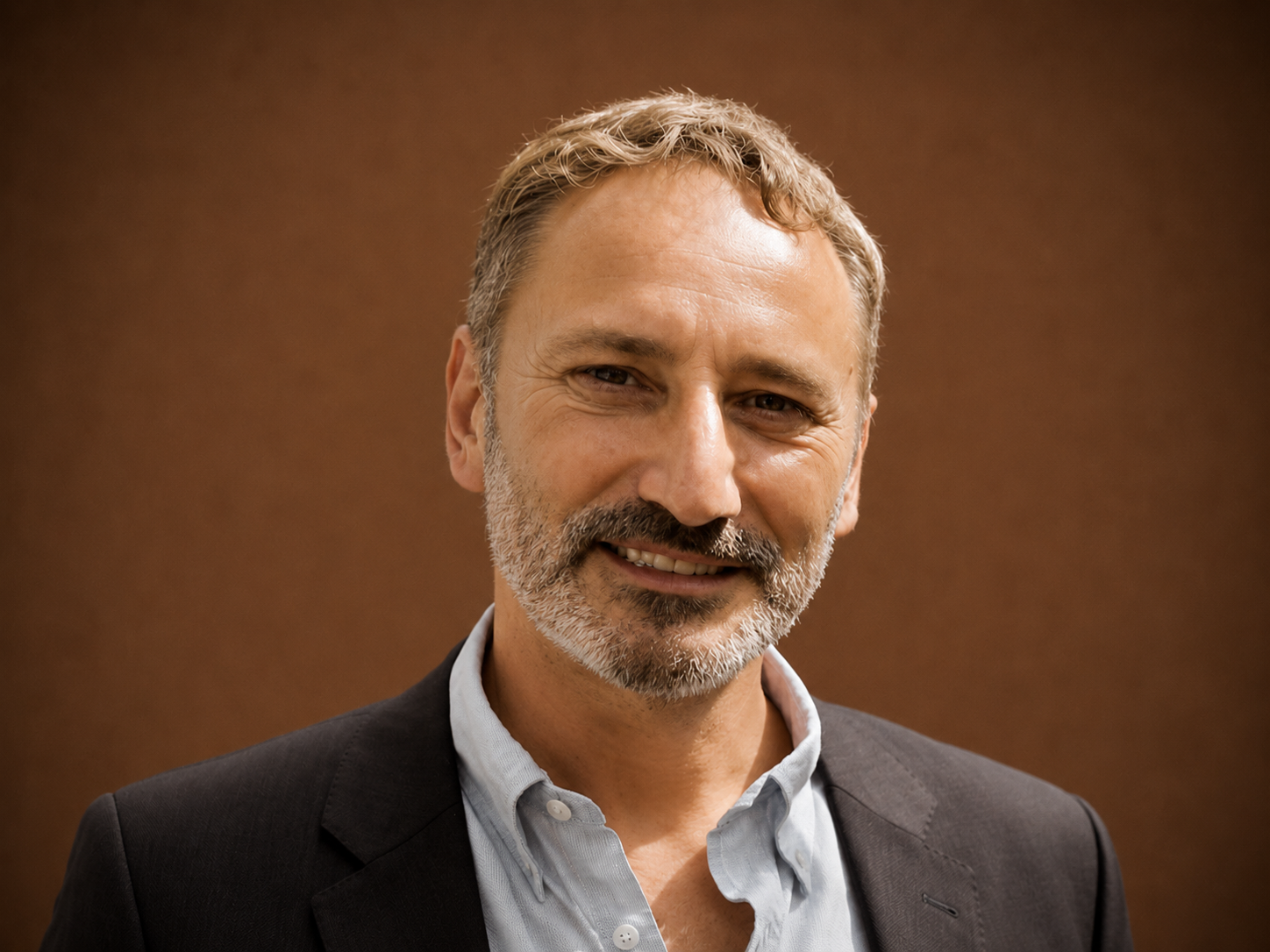 A smiling middle-aged man with gray hair and beard, wearing a dark blazer and light blue shirt, standing against a brown background.