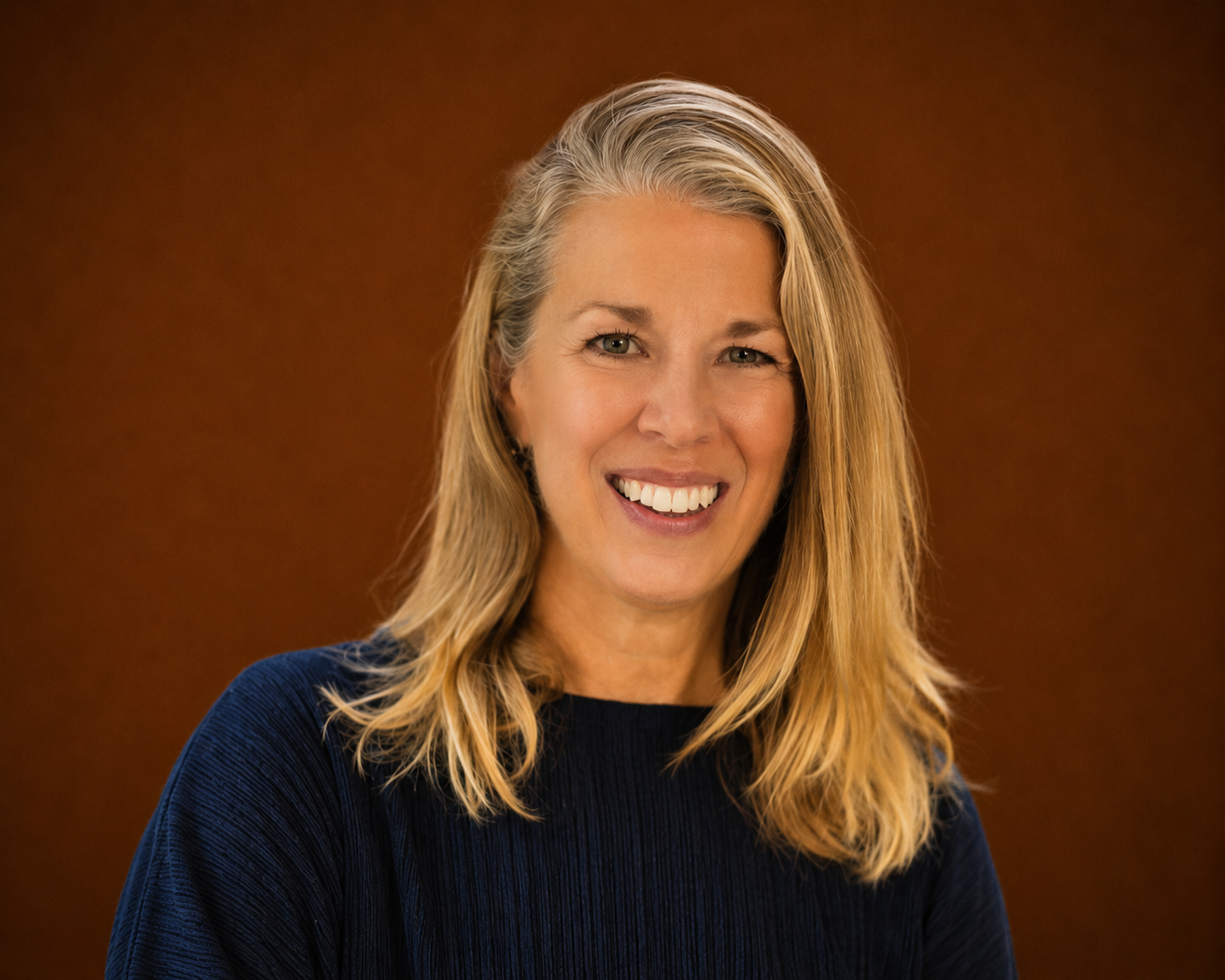 Portrait of a woman with blonde hair smiling, wearing a dark blue top, against a brown background.