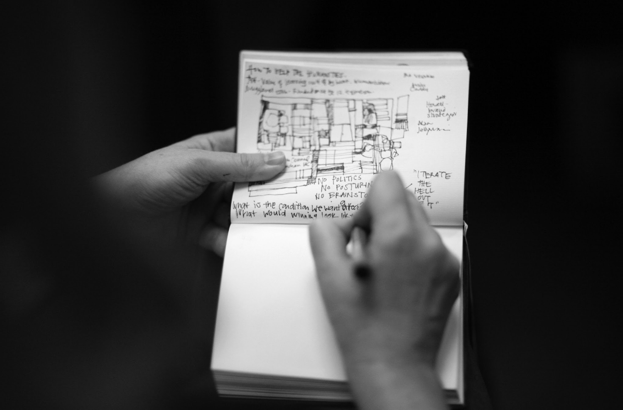 A person holding a sketchbook with handwritten notes and a drawn illustration, writing in a black notebook against a dark background.
