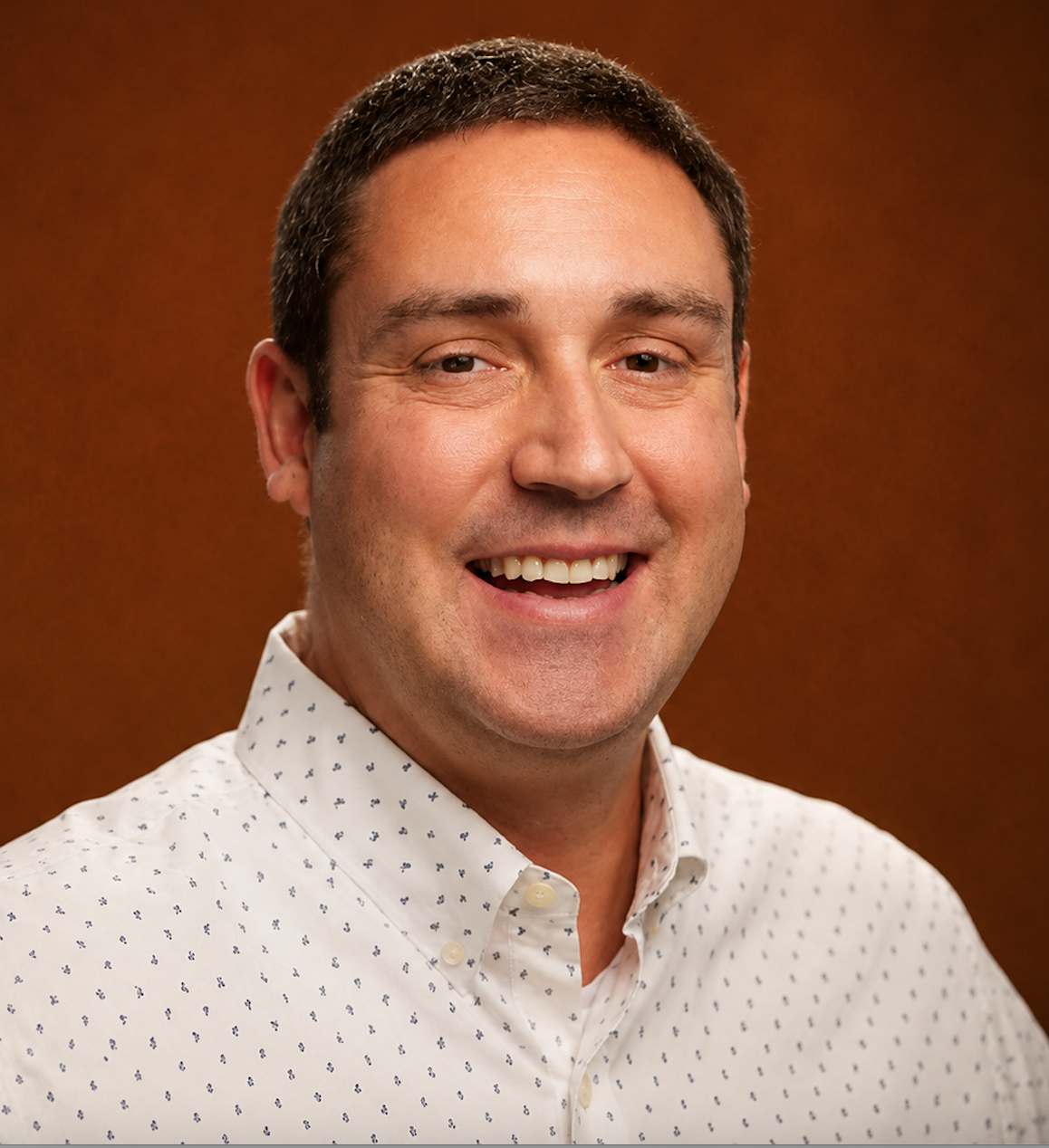Smiling man with short dark hair wearing a white collared shirt with small dark patterns, posing in front of a brown background.