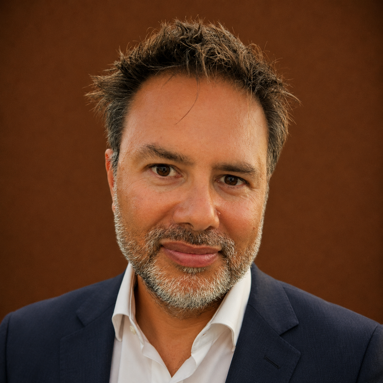 Close-up portrait of a middle-aged man with salt-and-pepper hair and beard, wearing a navy suit and white shirt, smiling slightly against a brown background.