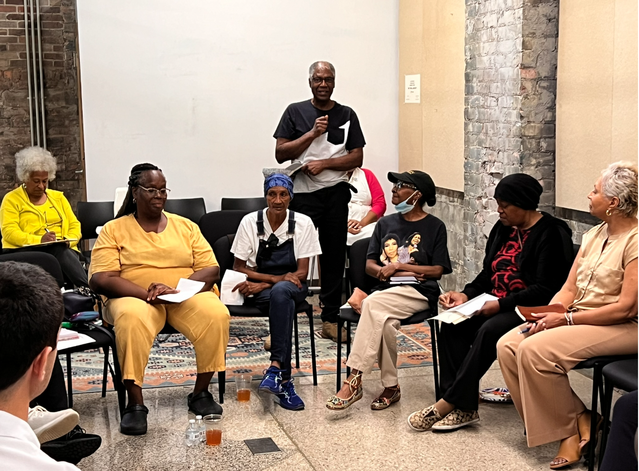 A group of diverse women seated in a semi-circle, participating in a discussion or meeting, with a man standing and speaking in the center.