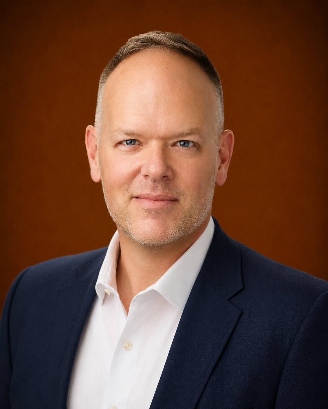 Professional headshot of a middle-aged man with short light brown hair, blue eyes, and a light beard, wearing a navy blazer and white shirt against a brown gradient background.