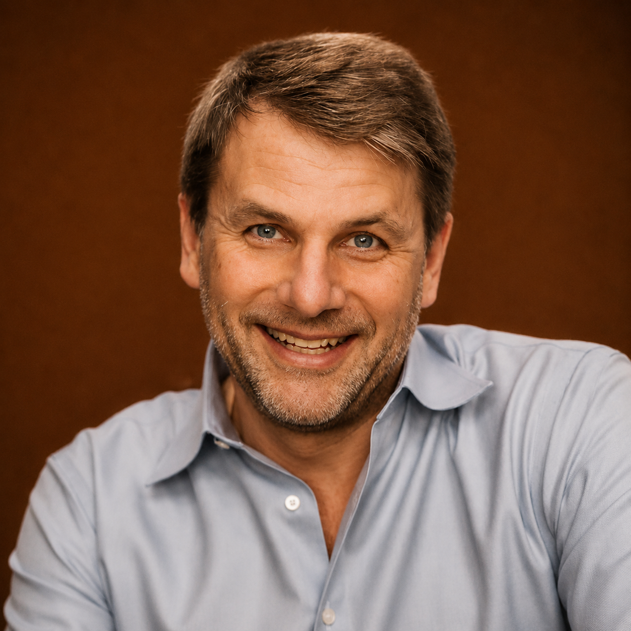 A smiling man with gray hair and a beard, wearing a light blue button-up shirt, looking directly at the camera.