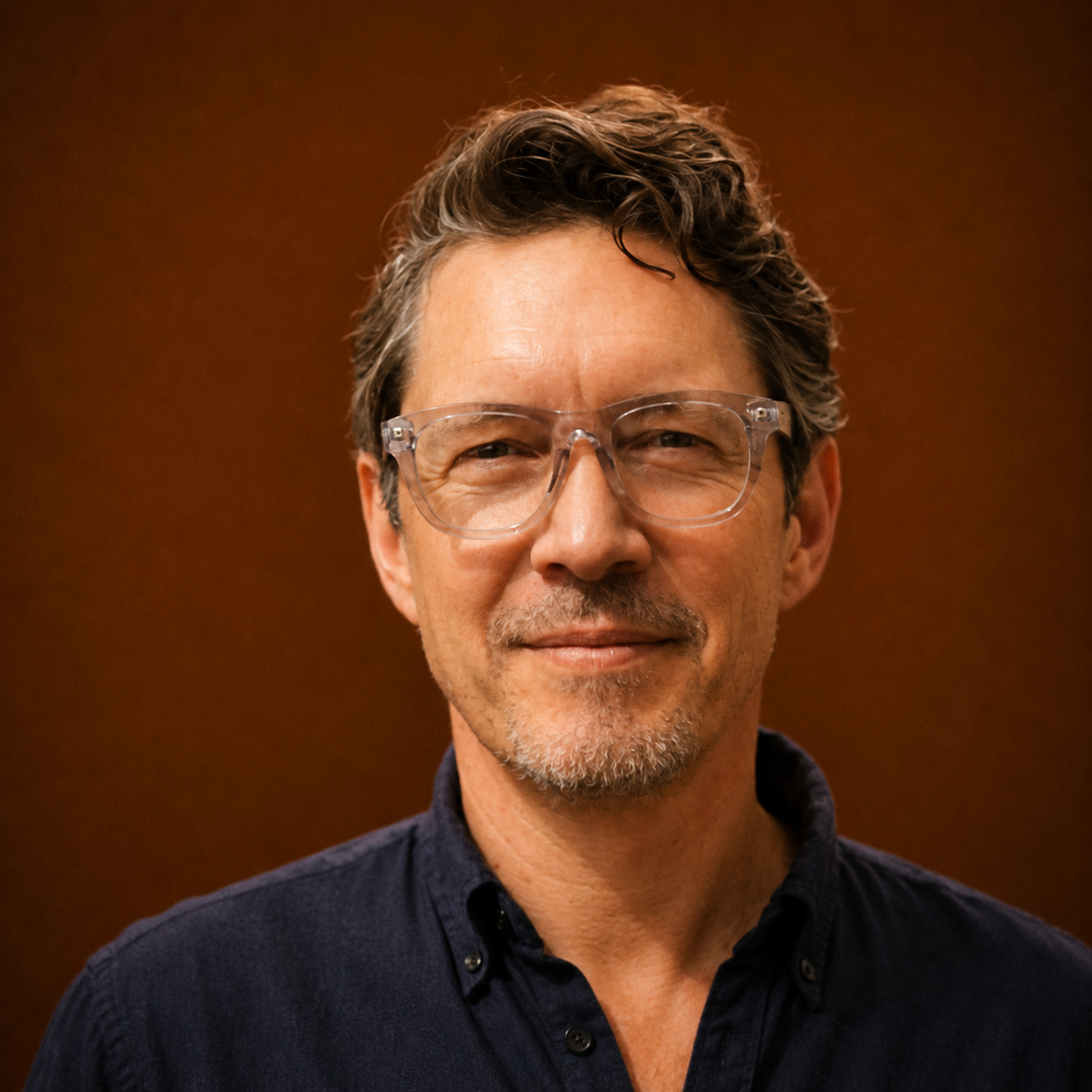 A smiling middle-aged man with short, wavy brown hair, wearing clear eyeglasses and a dark shirt, standing against a brown background.