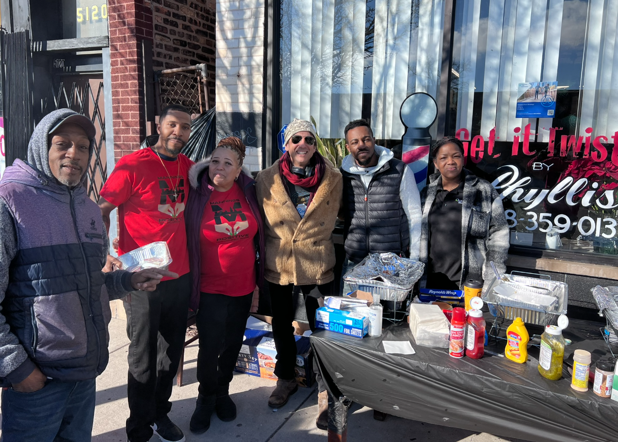 Group of six people standing at a street-side barbecue stand with food and condiments on a table, outdoors on a sunny day.