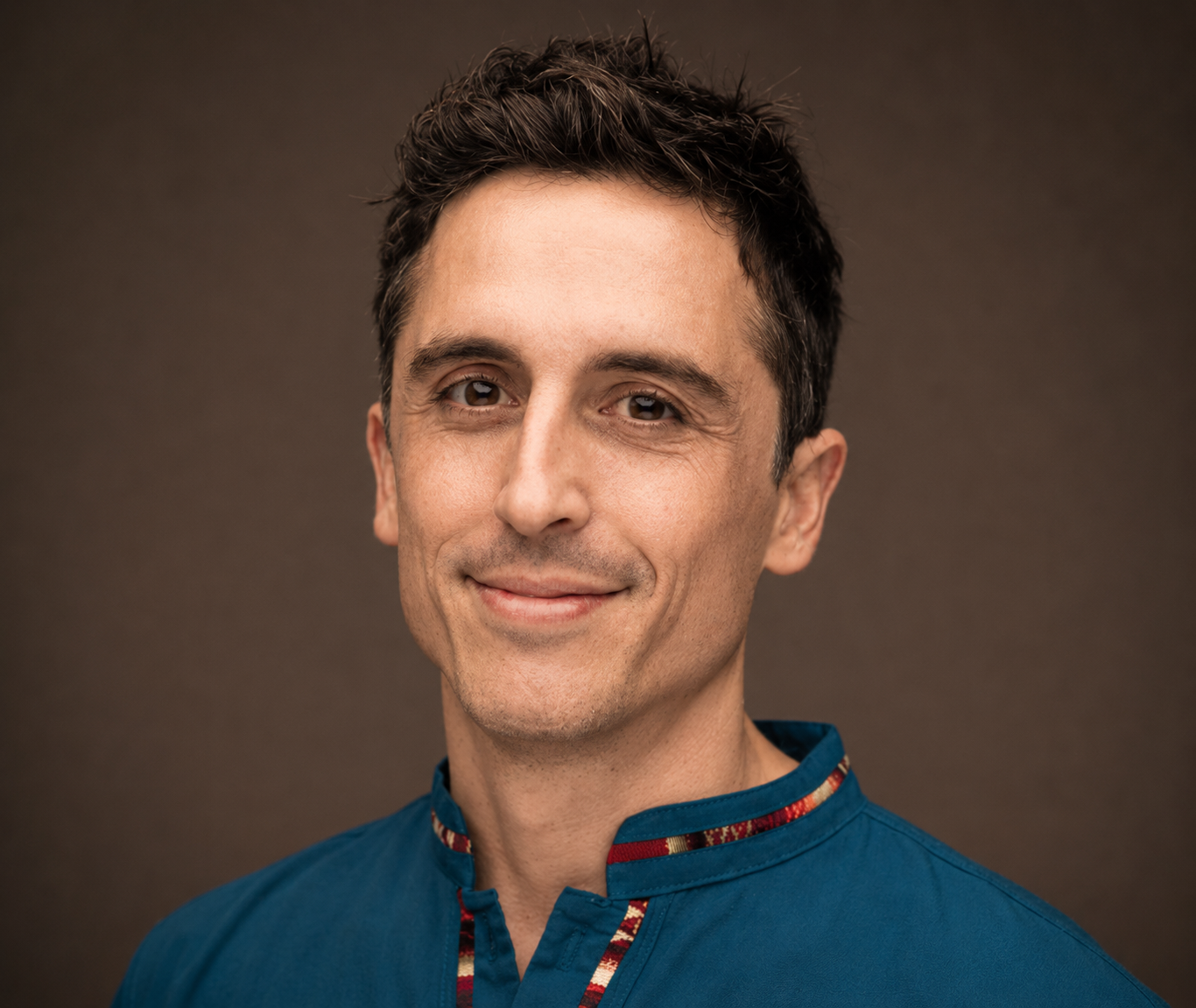 Portrait of a man with short, dark, curly hair wearing a traditional blue Asian garment with red and gold trim, smiling softly, against a plain brown background.