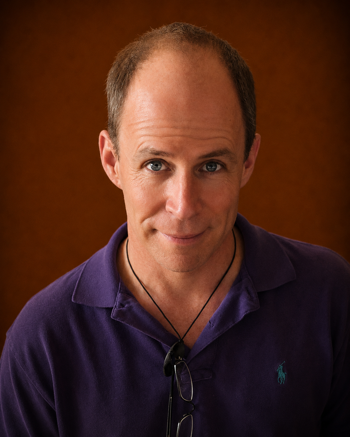 A man with short, light brown hair and blue eyes, wearing a purple Polo Ralph Lauren shirt, looking directly at the camera with a slight smile and glasses hanging from his shirt collar, against a brown background.
