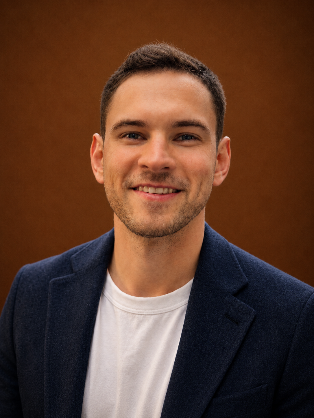Portrait of a young man with short brown hair, blue eyes, and light stubble, smiling, wearing a white T-shirt and navy blazer, against a brown background.