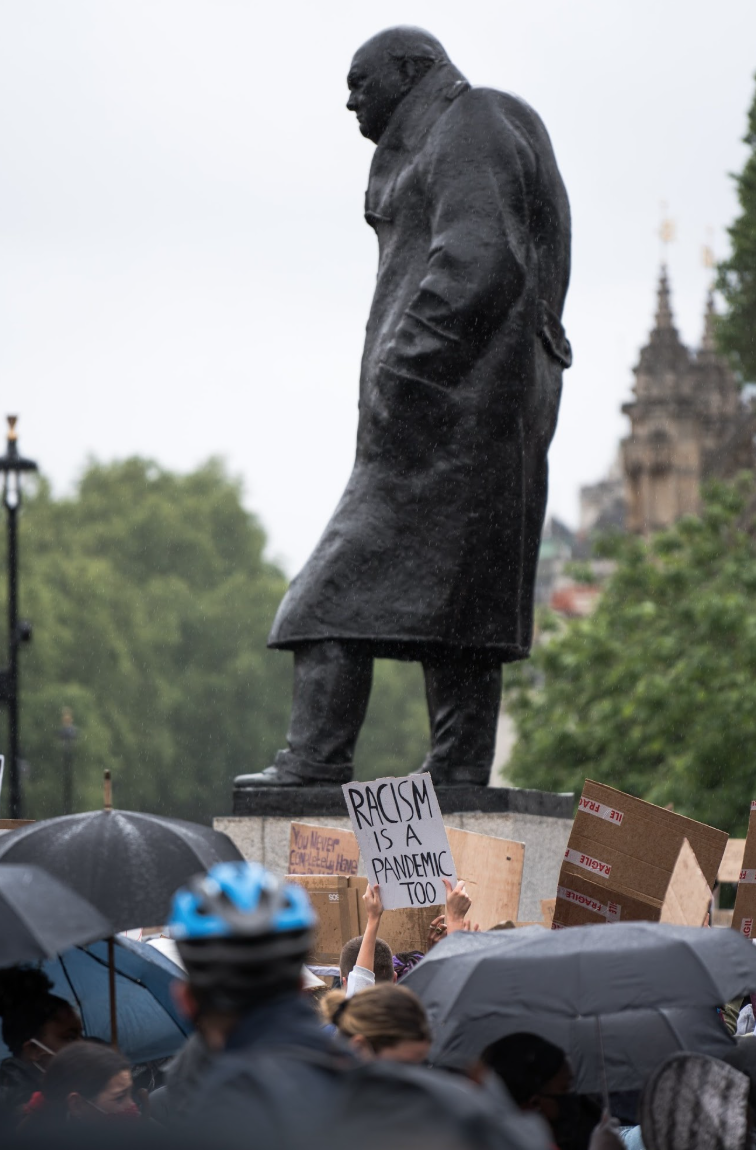 A statue of a man stands behind a crowd of protesters holding signs and umbrellas during a rainy day. One protester’s sign reads "RACISM IS A PANDEMIC TOO."