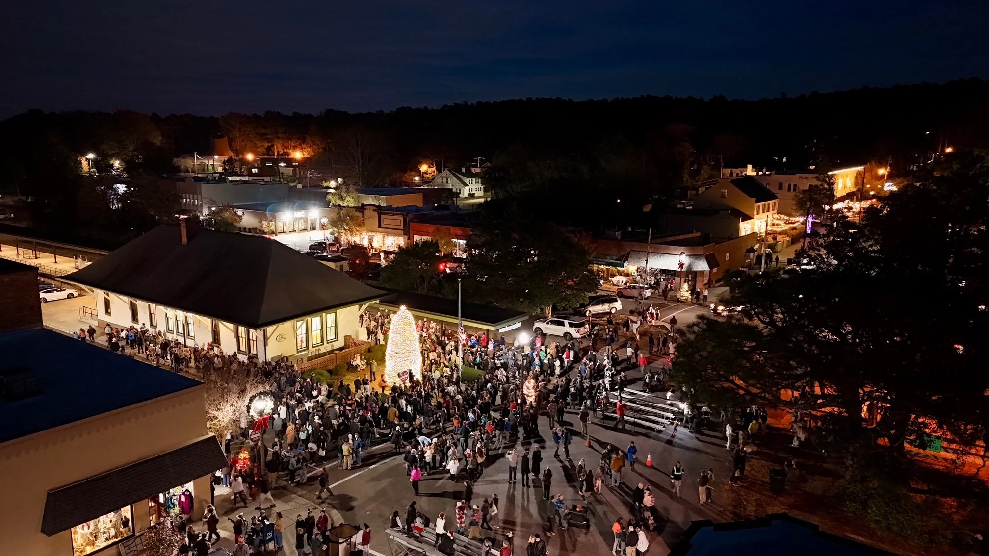 Nighttime scene of a lively street decorated for the holidays, with a large crowd gathered around a lit Christmas tree, and buildings illuminated in the background.