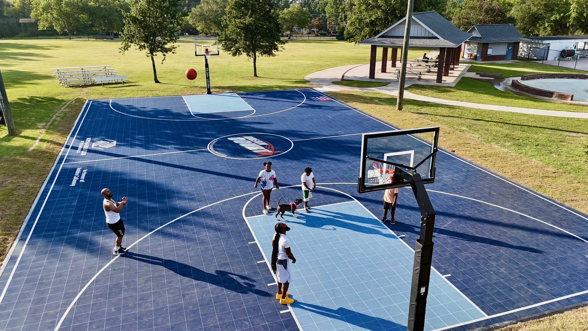 People playing basketball on an outdoor court with a solar panel surface, surrounded by grass, trees, and a pavilion.