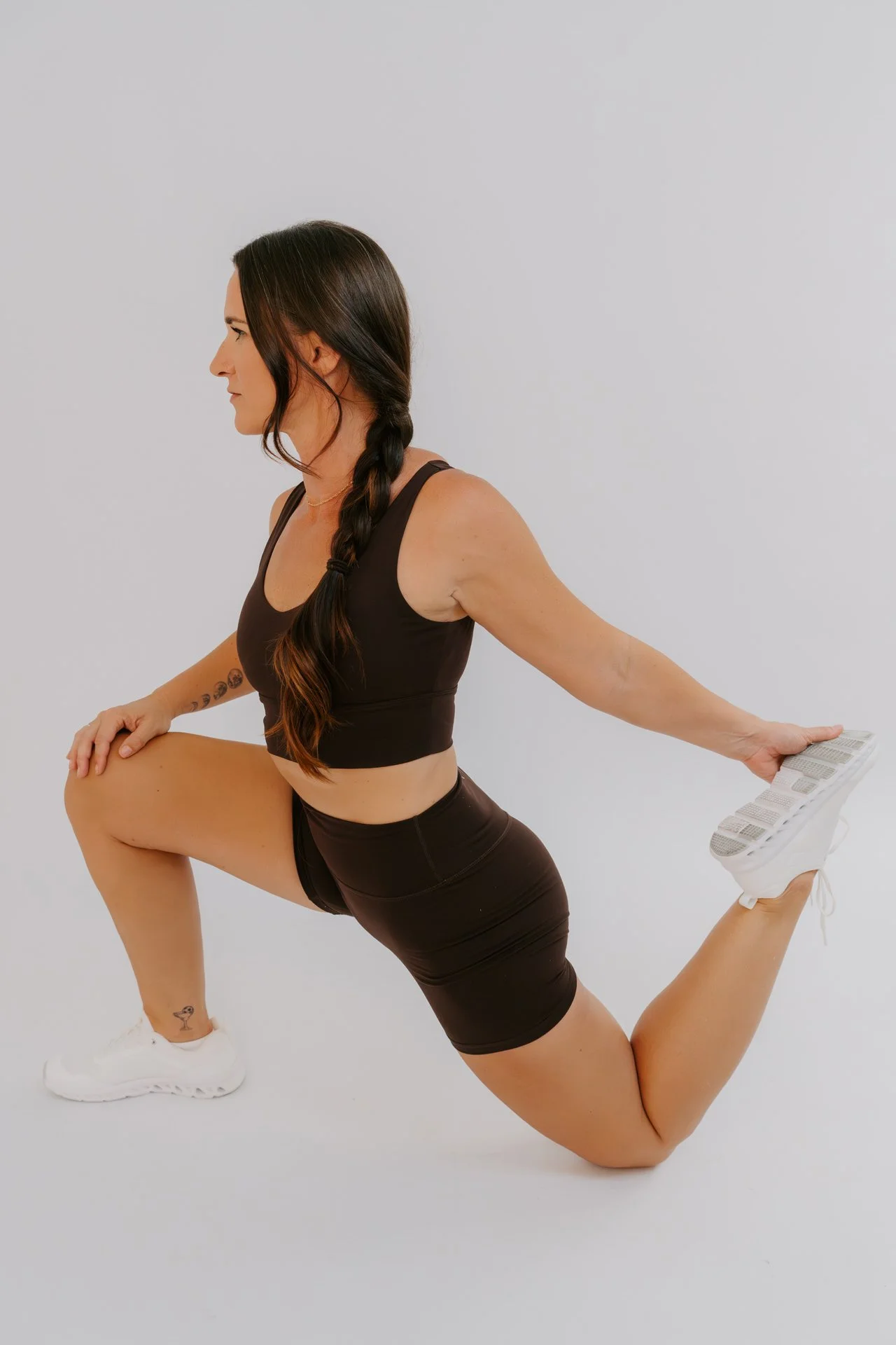 A fitness model with long brown hair tied in a braid doing a low lunge stretch wearing a matching brown activewear set and white sneakers