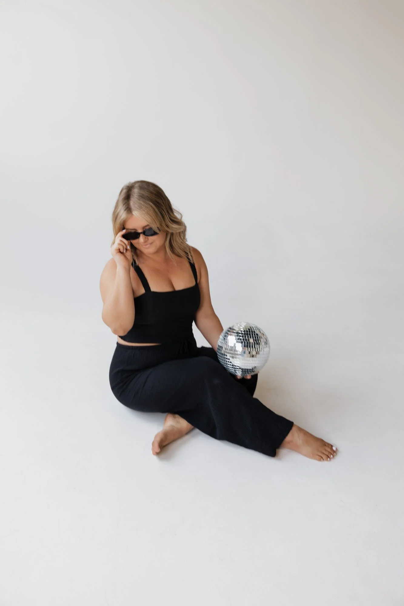 Alaina, founder of Alaina Marie Brand Photography, wearing a matching black linen set tilts her sunglasses as she looks down while holding a disco ball in a studio photoshoot