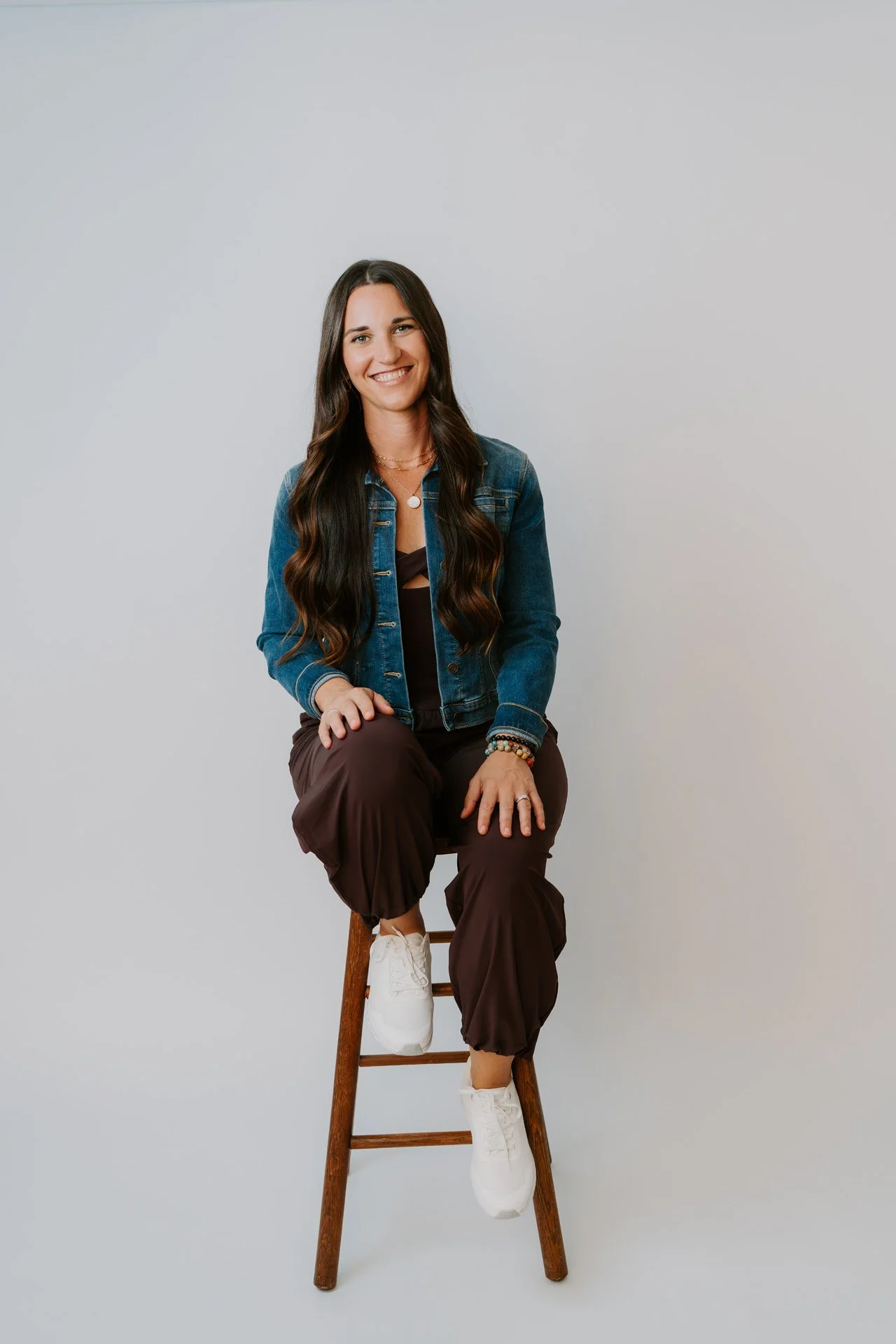 A woman with long brown hair wearing a brown romper and a jean jacket sits on a brown stool in a white studio setting while smiling at the camera