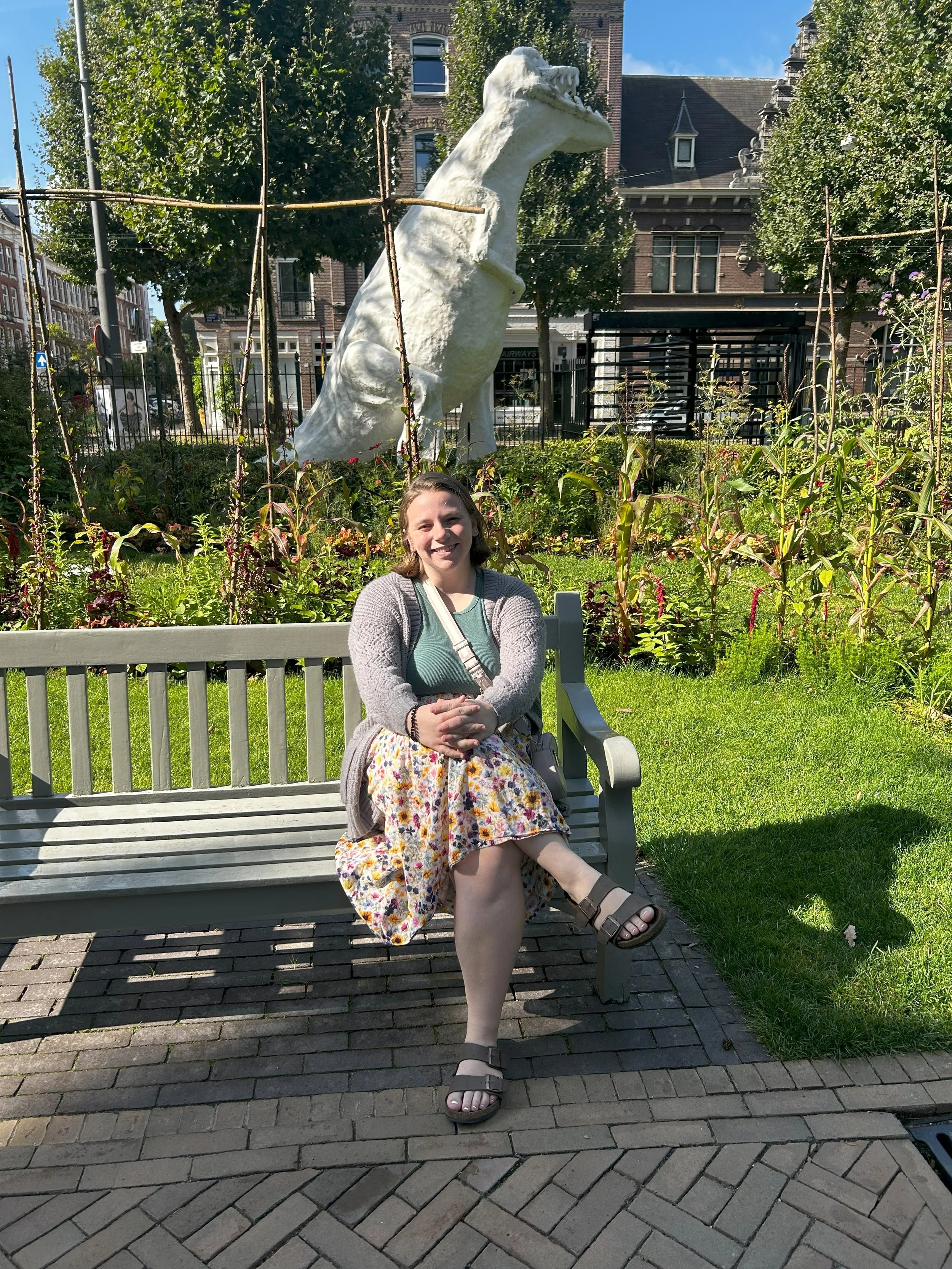 A woman sitting on a park bench with a garden and a large white sculpture of a horse's head in the background.