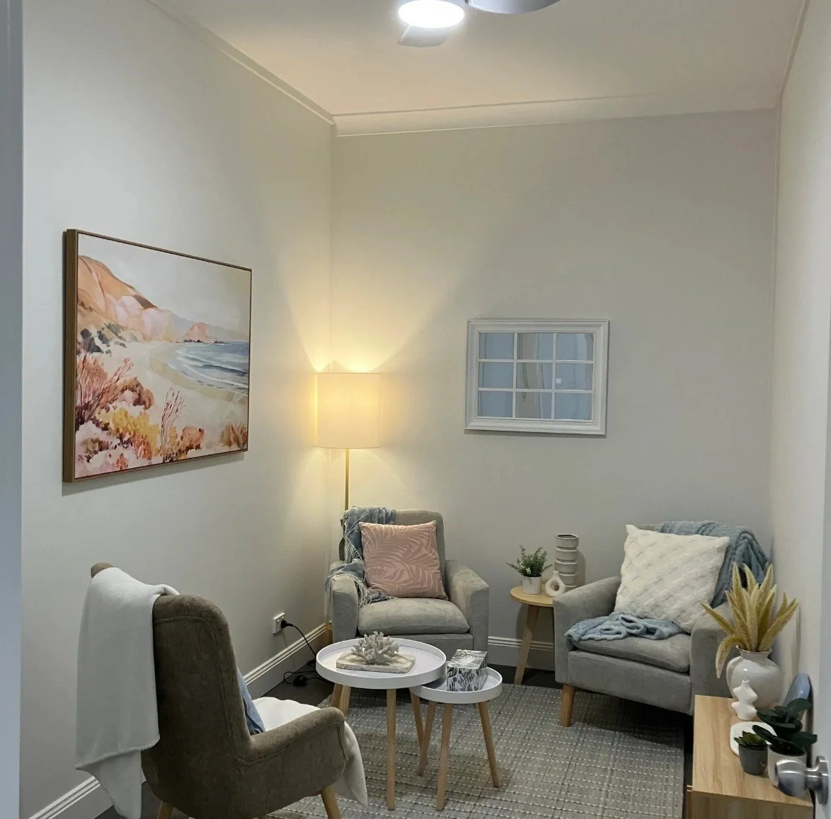 Cozy therapy room with neutral-colored furniture, a lamp, wall art, small side tables, and decorative plants.