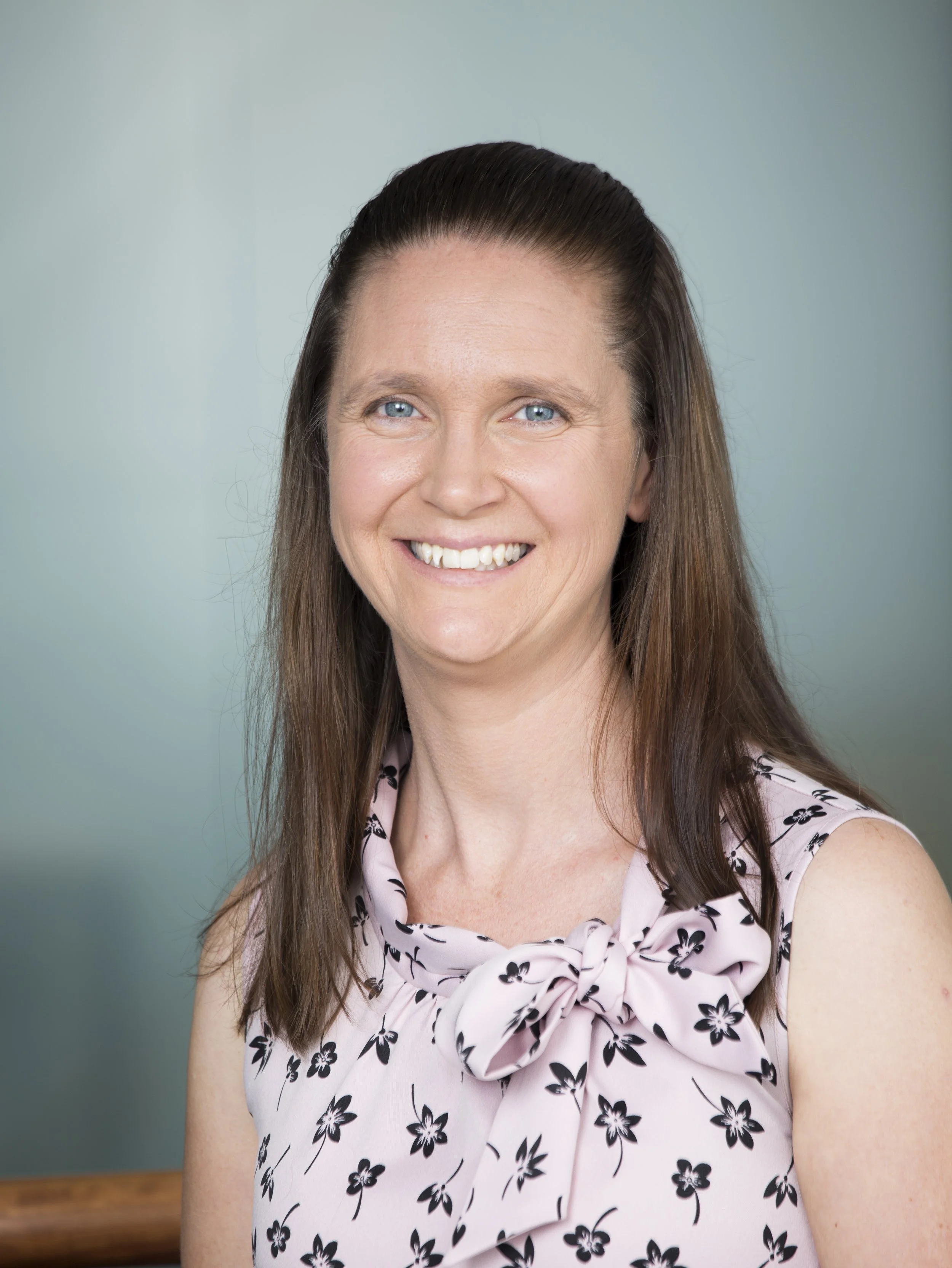 Close-up of a smiling woman with long brown hair, blue eyes, wearing a pink sleeveless blouse with black floral pattern and a knot detail at the neckline, against a light blue background.